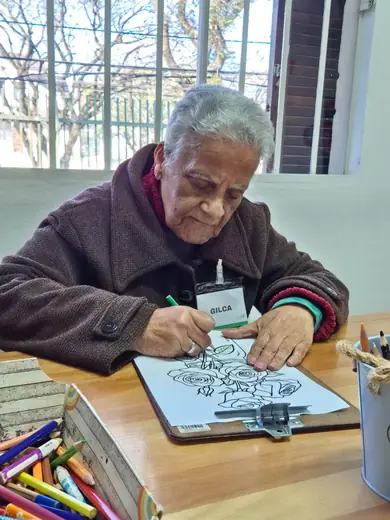 Senhor de casaco vinho desenhando a caneta numa prancheta, com potes de canetinhas coloridas sobre a mesa