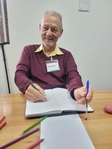 Senhor sorrindo à mesa, com o caderno de atividades aberto e a caneta na mão