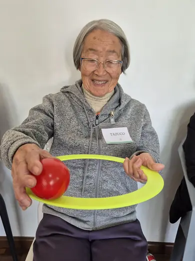 Senhora sentada esticando os braços à frente, com uma bola vermelha numa mão e um arco na outra