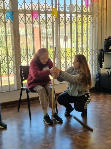 Senhora sentada segurando um bastão de ginástica, com a educadora agachada ao lado orientando o movimento