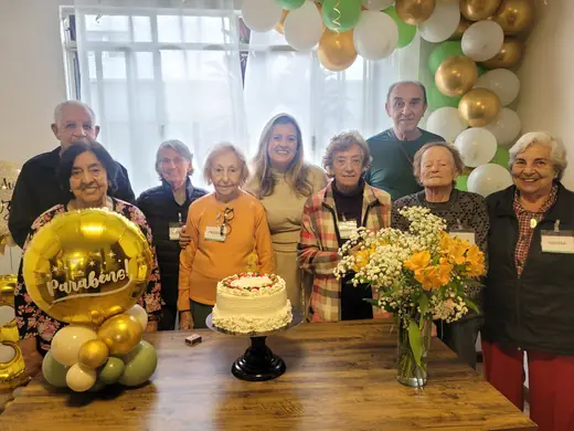 Idosos e equipe ao redor do bolo de aniversário, com balões dourados e verdes ao fundo