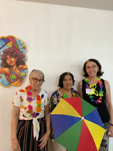 Três senhoras com colar de flores segurando juntas uma sombrinha colorida de frevo