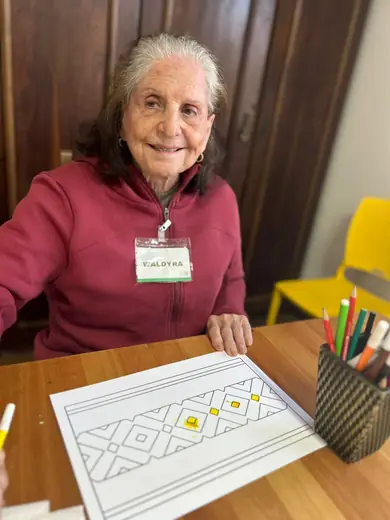Senhora sorrindo à mesa, com um desenho geométrico para pintar e um pote de canetas ao lado